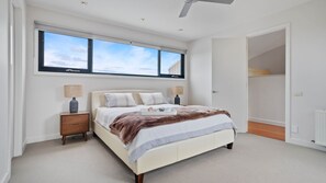 A modern bedroom with a large bed, two bedside tables, a ceiling fan, and a view of the sky through a large window.