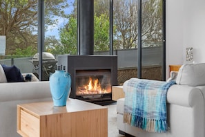 A modern living room with a fireplace, a sofa, a coffee table, and a view of trees through large windows. - St Johns Wood Treehouse (Blairgowrie)