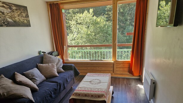 Une pièce chaleureuse avec un canapé bleu foncé, une petite table en bois recouverte d’une nappe à motifs floraux et une vue sur un balcon doté d’une rambarde verte.