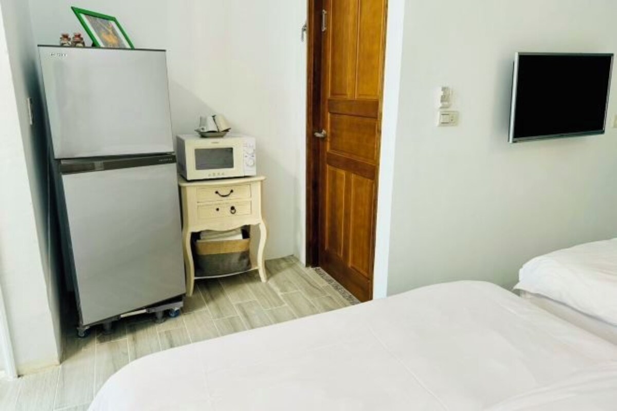A hotel room with a bed, a small bedside table with a microwave, a refrigerator, and a wooden door.