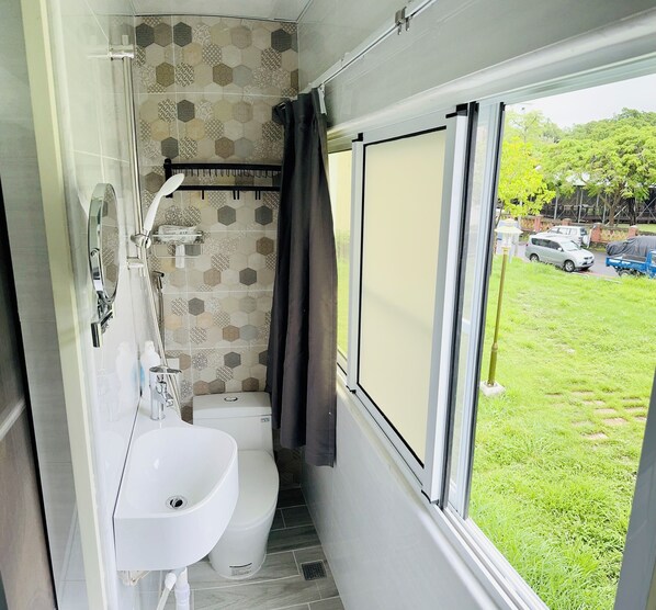 Baño con inodoro, lavamanos y ducha. Tiene una ventana con vista a un área verde y coches aparcados afuera.