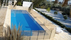 En rektangulär swimmingpool med klart, blått vatten, omgiven av en kakelbeklädd terrass och ett område med grus och små växter.