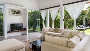 A modern living room with a large TV, comfortable sofas, and a view of a lush garden. - Villa Alice Dua by Villa Finder (Seminyak)