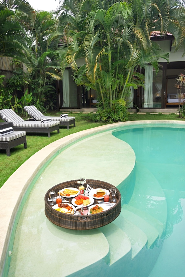 A pool with a floating dining setup, lounge chairs, and lush greenery.
