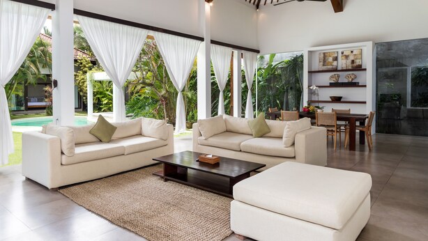 A spacious living area with two sofas, a coffee table, and a dining table with chairs. There is a large window with curtains, a ceiling fan, and a view of a pool and greenery outside. - Villa Alice Dua by Villa Finder (Seminyak)