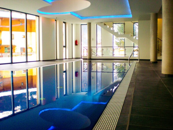 A spacious indoor swimming pool with clear glass walls, a central water feature, and a staircase leading to an upper level.
