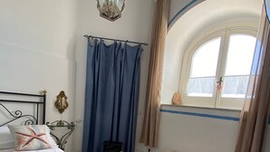 Una habitación con una cama, un pequeño banco, un ventanal con cortinas y un suelo estampado.