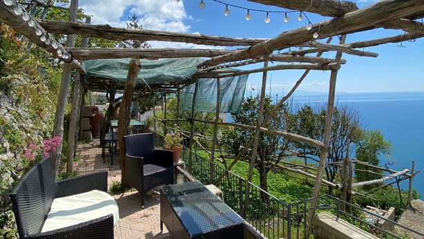 Un patio exterior con mobiliario de mimbre, una pérgola con luces de cuerda y vistas al mar.