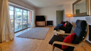 A spacious living room with a large sliding glass door leading to a balcony, a flat-screen TV, a wooden floor, and a cozy seating area with a sofa and chairs. - Cosy House in Eigionfa (Menai Bridge)