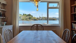 A dining area with a table and chairs, a window with a view of a lake and trees, and a bookshelf on the right. - Cosy House in Eigionfa (Menai Bridge)
