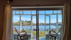 A room with a large sliding glass door leading to a balcony with two wicker chairs and a view of a lake and trees. - Cosy House in Eigionfa (Menai Bridge)