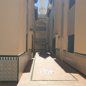 Un couloir aux murs décorés de motifs et au sol carrelé, qui mène à une cour d’où l’on peut admirer le ciel.