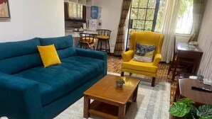 Ein Wohnzimmer mit blauem Sofa, gelbem Sessel, Couchtisch aus Holz und einem Essbereich mit Tisch und Stühlen.