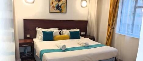 Habitación de hotel con cama, mesita de noche, ventilador de techo, un cuadro en la pared y ventana con cortinas.