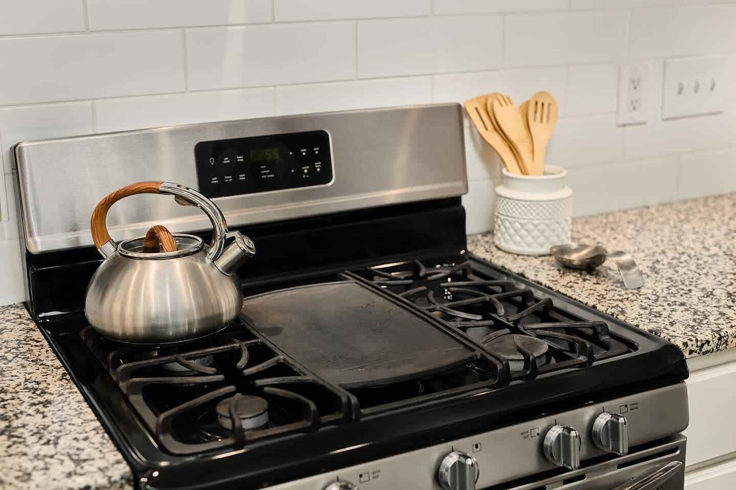 A kitchen with a stainless steel kettle on the stove, a digital control panel, and wooden utensils in a holder.