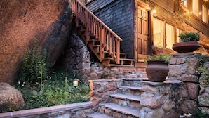 Una escalera de piedra que conduce a una puerta de madera, rodeada de vegetación y una gran formación rocosa.