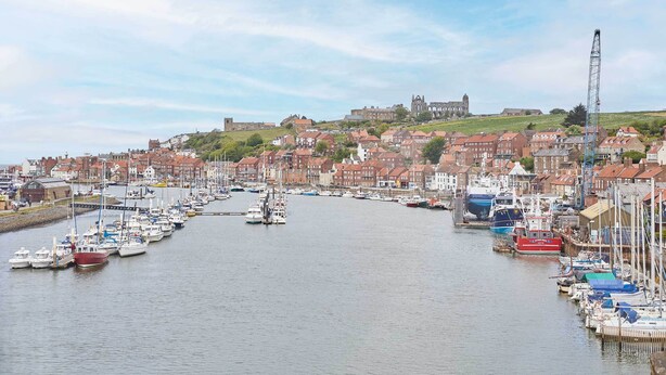 A harbor with numerous boats docked along the waterfront, surrounded by historic buildings and a prominent castle on a hill in the background.