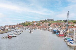 Um porto com inúmeros barcos atracados ao longo da orla, cercado por edifícios históricos e um castelo imponente numa colina ao fundo.