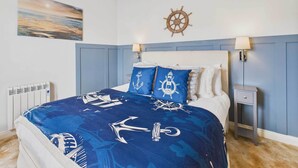 A bedroom with a bed, two nightstands, a wall-mounted ship's wheel, and a painting of a sunset.