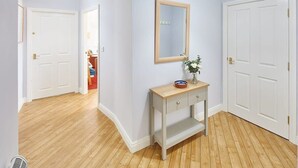 A hallway with a wooden floor, white walls, and a light blue door. There is a small table with a mirror and a vase with flowers.