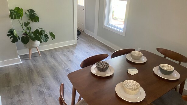 Un comedor con una mesa de madera dispuesta para cuatro personas, rodeada de sillas. Se ve una planta en maceta en la esquina.