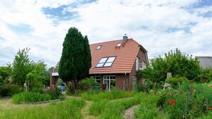 A house with a red-tiled roof, a large tree, and a garden with various plants and flowers. - Centrally located, quiet apartment with terrace and garden (Teschenhagen Sehlen)
