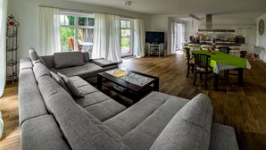 A modern living room with a sectional sofa, a dining table with chairs, a kitchen area, and large windows with curtains. - Centrally located, quiet apartment with terrace and garden (Teschenhagen Sehlen)