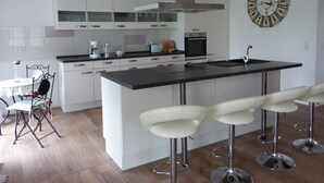 A modern kitchen with white cabinetry, a central island with bar stools, a built-in oven, and a wall clock. - Centrally located, quiet apartment with terrace and garden (Teschenhagen Sehlen)