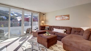 Ein Wohnzimmer mit einem braunen Ecksofa, einem Couchtisch mit Pflanze und einem großen Fenster mit Blick auf eine verschneite Landschaft.