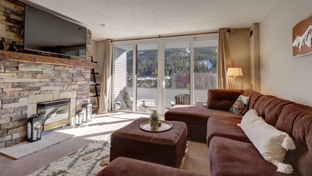 Ein Wohnzimmer mit Kamin aus Stein, einem großen Flachbildfernseher, einem braunen Ledersofa und Blick auf eine Berglandschaft durch große Fenster.