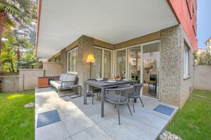 A patio with a table and chairs, a lamp, and a view of the interior through glass doors. - Res. San Marco 1 With Private Garden (Lugano)