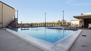 Une piscine rectangulaire bordée de bleu, entourée d’une terrasse en béton. On y trouve plusieurs chaises longues et un petit escalier menant à un niveau supérieur.