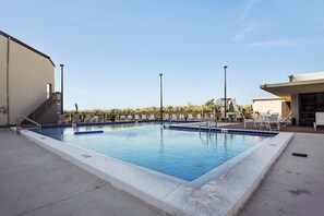 Una piscina rettangolare con bordo blu, circondata da una terrazza in cemento. Si vedono diversi lettini e una piccola scala che conduce a un livello superiore.
