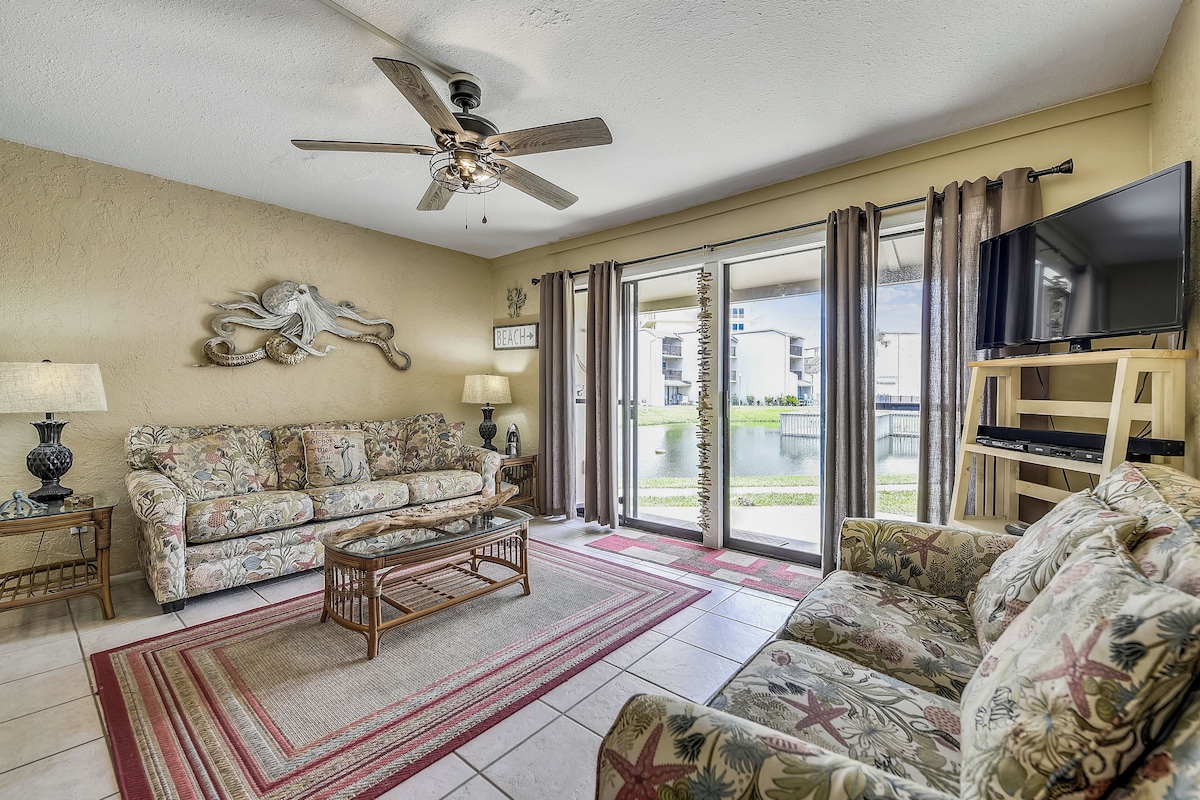 A living room with a ceiling fan, a sofa, a coffee table, a TV on a stand, and a view of the outside through large windows.