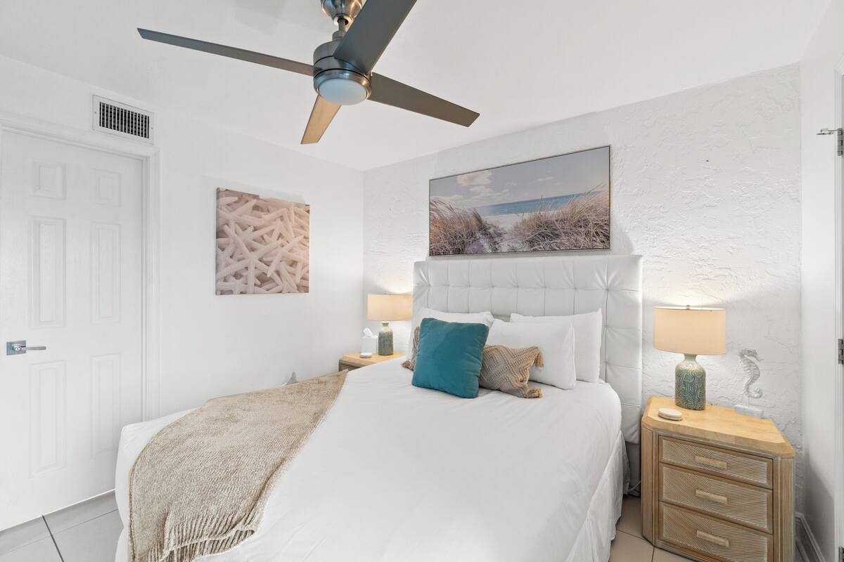 Une chambre à coucher comprenant un lit, un ventilateur de plafond, une table de chevet avec une lampe et un tableau accroché au mur.