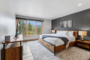 A bedroom with a large bed, a desk, a chair, a window with a view of trees and mountains, and a wall-mounted artwork.