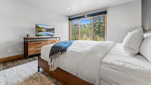 Une chambre à coucher avec un lit, un téléviseur posé sur une commode en bois et une grande fenêtre donnant sur des arbres et des montagnes.