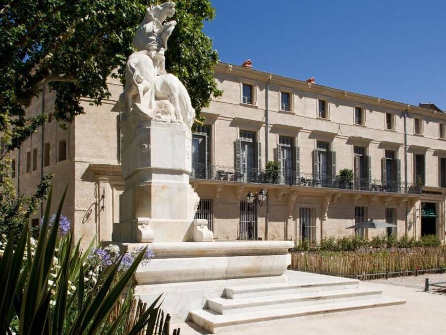 Un edifici amb façana clàssica, una estàtua central i un pati amb escales cap a l'entrada.