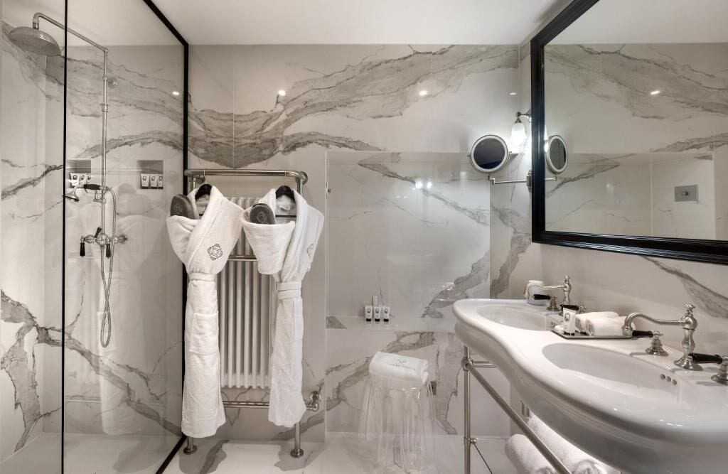 A modern bathroom with a glass-enclosed shower, a marble-tiled wall, a large mirror, and a white sink with a vanity.