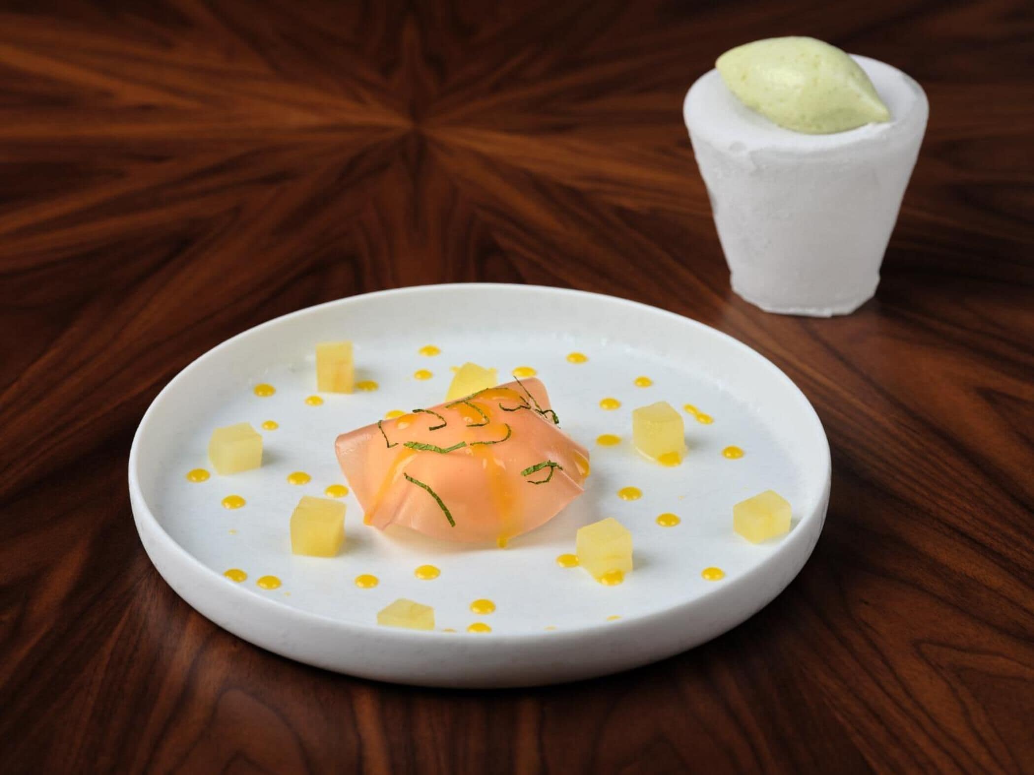 Um prato branco com prato de salmão, cubos de manteiga e um pequeno copo de molho verde sobre uma mesa de madeira.