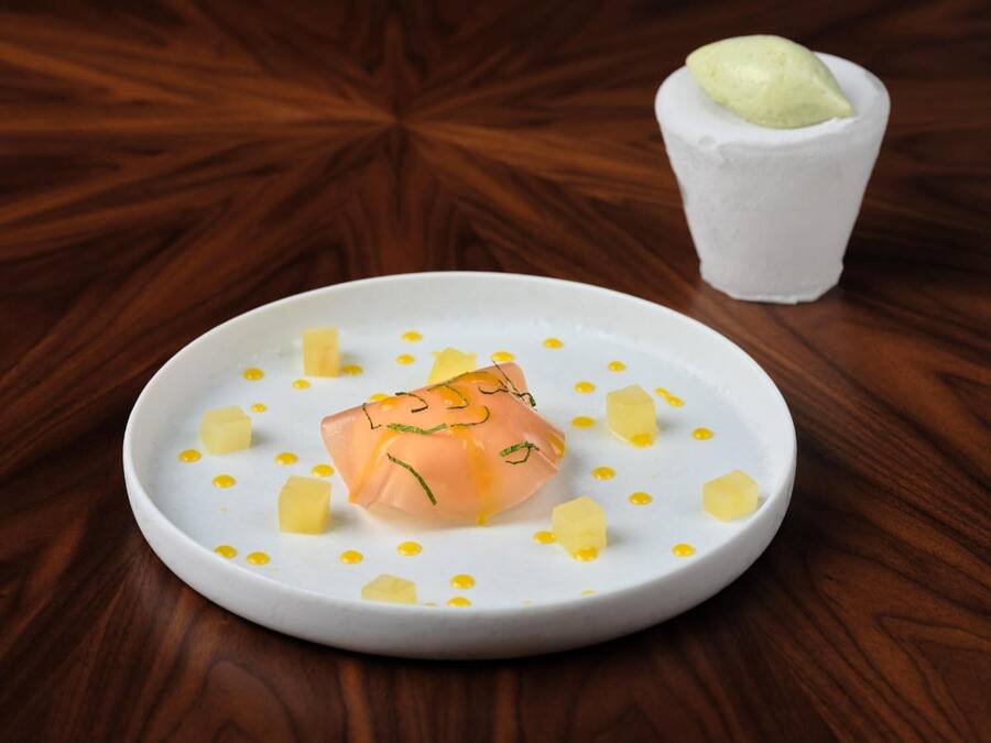 Un plato blanco con un plato de salmón, cubos de mantequilla y un vaso pequeño de salsa verde sobre una mesa de madera.