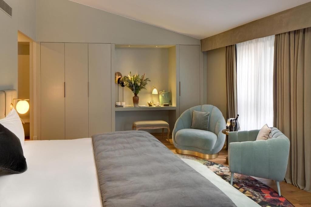 A hotel room with a large bed, a seating area with two armchairs, a small table, and a desk with a lamp and decorative items.