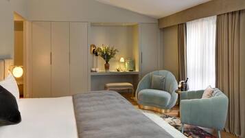 A hotel room with a large bed, a seating area with two armchairs, a small table, and a desk with a lamp and decorative items.