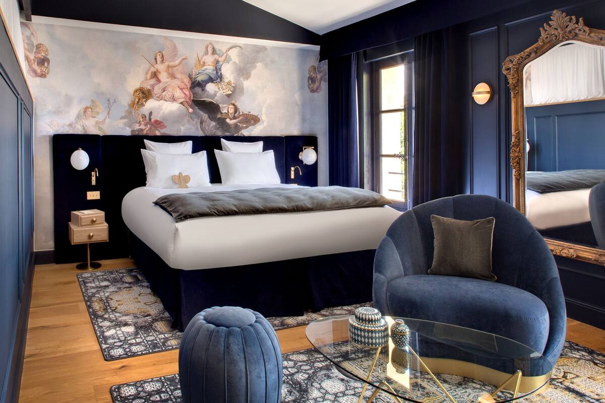 A modern bedroom with a large bed, a round armchair, a glass coffee table, and a decorative mirror.
