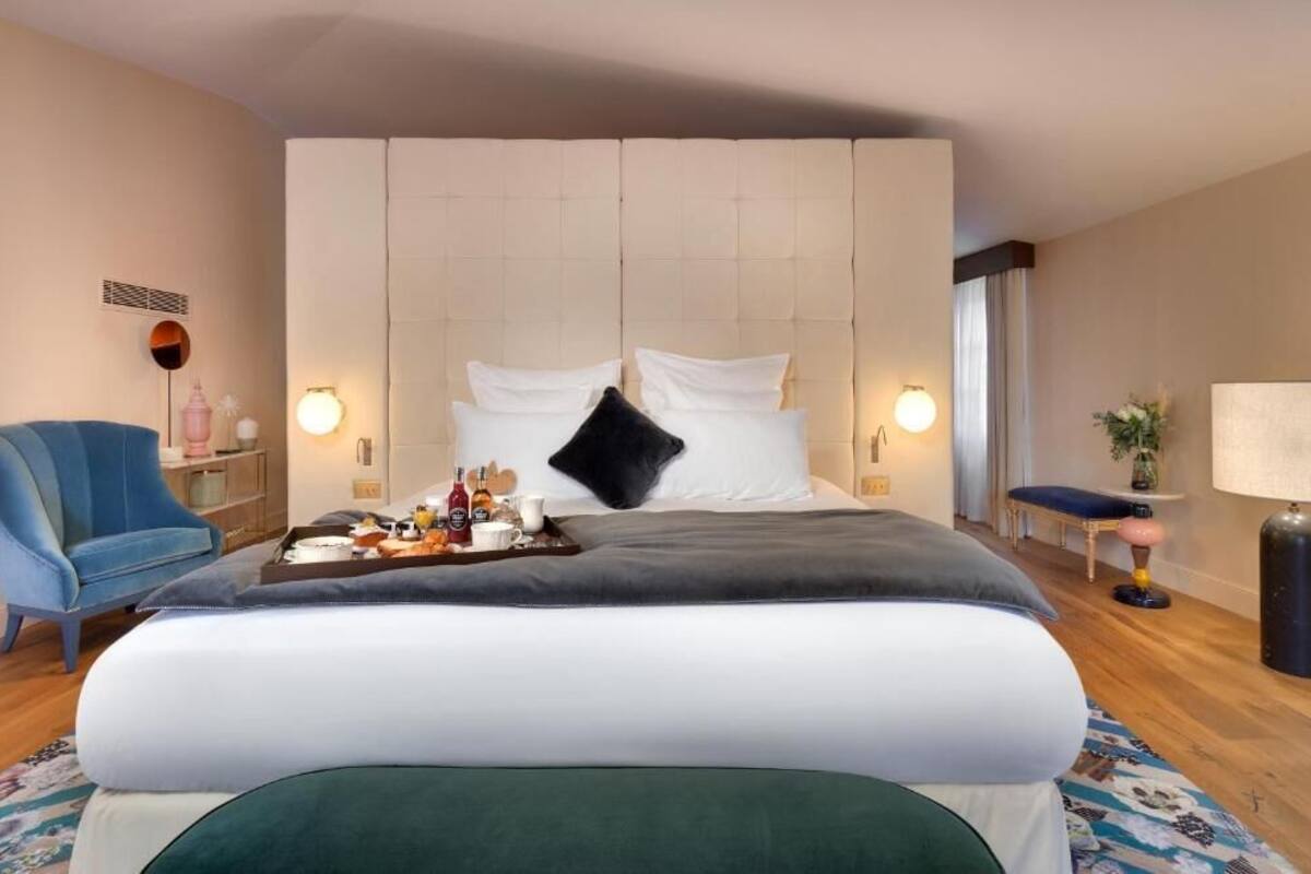 A hotel room with a large bed, a blue armchair, a bedside table with a tray of items, and a lamp.