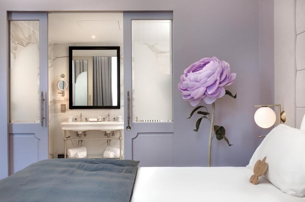 Un dormitorio con una cama grande, una mesita de noche, un espejo en la pared, una tocador con lavabo y una decoración de una gran flor morada.