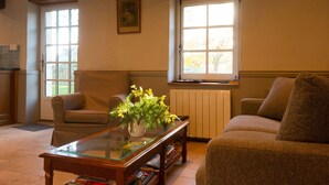 Een woonkamer met een glazen salontafel, twee bruine fauteuils, een beige bank, een radiator en een raam met uitzicht op bomen.