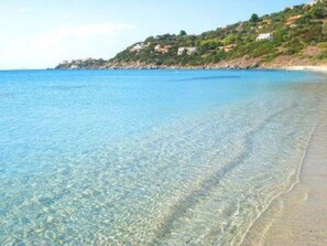 A clear coastal beach with visible underwater features and a hillside with buildings.