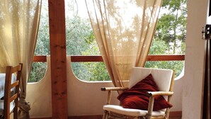 Una silla de madera con cojín, una mesa de madera y vistas a la vegetación a través de una ventana con cortinas de gasa.
