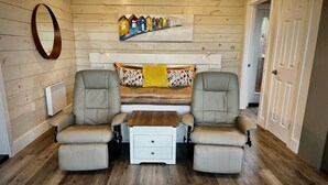 A room with wooden panel walls, a whiteboard, a painting of colorful houses, two beige armchairs, a small white table, and a wooden floor. - Whitecaps Cabana for Couples (Riverport)